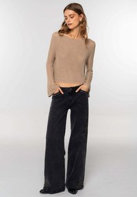 Top de punto beige de manga larga con mangas de campana, combinado con unos pantalones de mezclilla negros de pierna ancha de tiro alto, que presentan acentos de tachuelas a lo largo de los lados.