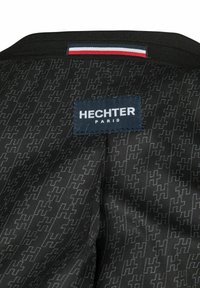 Zwarte stof met herhalend "H"-patroon, rechthoekig Hechter Paris-label in wit op marineblauwe achtergrond, en stoflabel met rode, witte en blauwe strepen.