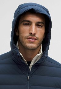 Sötétkék puffertakaró kapucnival, steppelt mintázattal, magas gallérral és cipzáras záródással. Az anyag simának és könnyűnek tűnik.