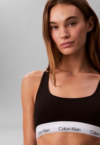 Jeune femme aux cheveux bruns raides portant un soutien-gorge de sport noir Calvin Klein sur un fond gris uni.