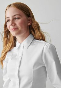 Camicia bianca con colletto classico, placket testurizzato e maniche a sbuffo, realizzata in tessuto liscio. Adatta per un look casual o formale.