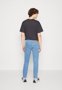 Camiseta negra, jeans de mezclilla azul claro con un parche de cuero marrón y zapatillas grises. Los jeans tienen un corte clásico y bolsillos traseros.