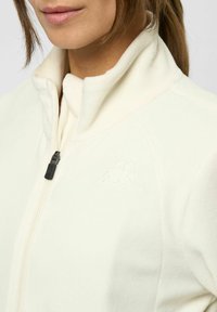 Valkoinen fleece-takki, jossa on korkea kaulus ja vetoketjukiinnitys. Hellävaraista logoa vastaavassa värissä rinnassa. Pehmeä tekstuuri, istuva malli.