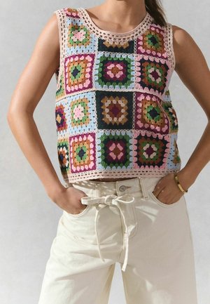 Femme portant un top sans manches en crochet avec des motifs carrés colorés et un pantalon taille haute écru avec ceinture nouée, mains dans les poches.