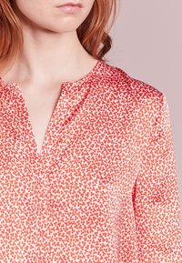 Rød bluse med blomsterprint i blød stof, med V-hals og subtil glans, præsenteret mod en lys pink baggrund.