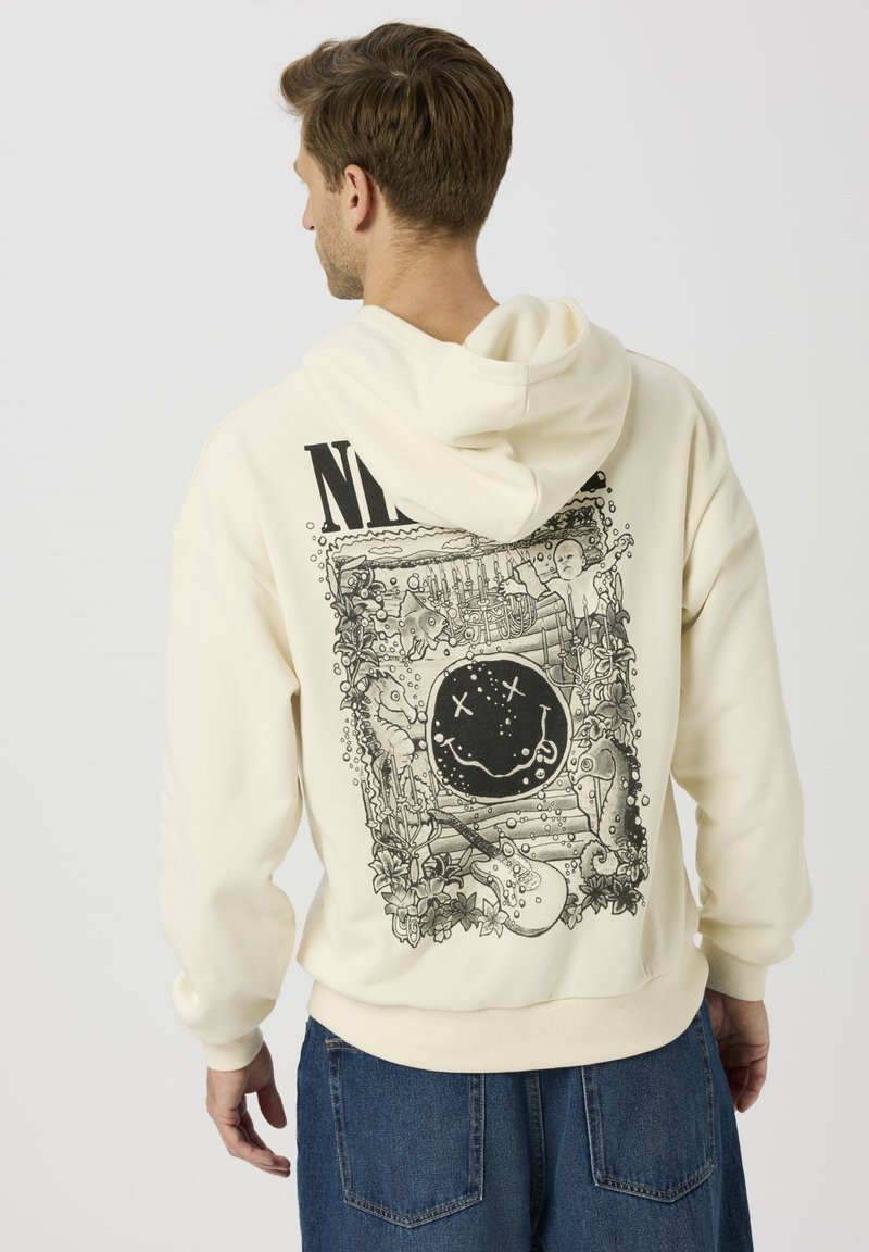 Beiger Hoodie mit großem schwarzem Grafikmotiv auf der Rückseite, das surreale Elemente wie ein Smiley-Gesicht, Pflanzen und ein Skelett zeigt, abgeschlossen mit gerippten Bündchen.