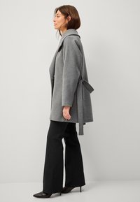 Grijze wollen jas met een brede kraag, knoopsluiting en zijstrikdetail. Gecombineerd met zwarte flared pants en puntige hakken.