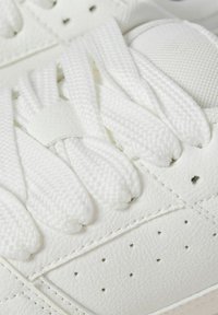 Weiße Sportschuhe mit einem strukturierten Obermaterial aus Leder, aufwendigen Nähten und flachen weißen Schnüssen, sowie kleinen Perforationen zur Atmungsaktivität.