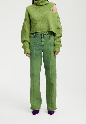 Persoon die een groene, korte gebreide trui met open schouders draagt, groene high-waisted jeans met acid-wash effect en paarse puntige schoenen, rechtstaand.