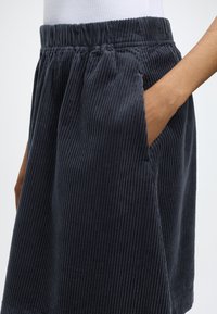 Personne portant une jupe en velours côtelé foncé avec une taille élastique, mettant une main dans la poche de la jupe, montrée de la taille vers le bas.