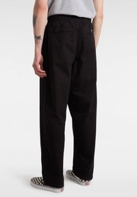 Personne portant un pantalon noir ample, un t-shirt gris et des chaussures slip-on à carreaux noirs et blancs, debout de dos au spectateur.