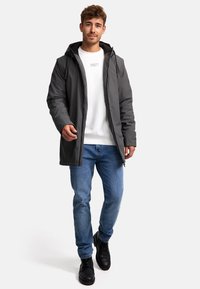 Doudoune grise avec une capuche, portée avec un sweat-shirt blanc et un jean bleu. Chaussures noires. Tenue décontractée en superpositions.