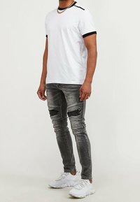 Camiseta blanca de manga corta con ribete negro, combinada con jeans ajustados grises desgastados y zapatillas deportivas blancas. Presenta un detalle de cadena en el escote.