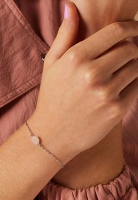 Zilveren kettingarmband met een lichtroze edelsteen kraal. Hand die de armband draagt, bevindt zich tegen een gestructureerde roze stofachtergrond.