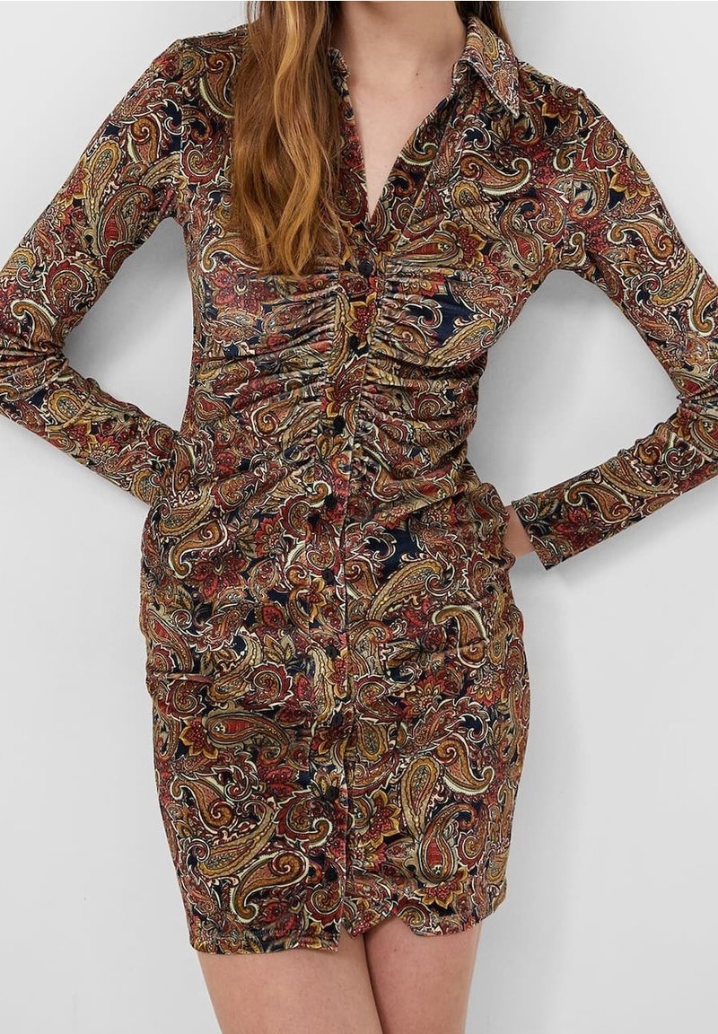 Femme portant une robe mini ajustée à manches longues, col, avec un motif cachemire multicolore en rouge, or, bleu marine et noir, mains sur les hanches.