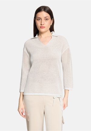 Femme portant un pull en maille blanche à manches 3/4 et un pantalon cargo beige, tournée vers l'avant sur un fond uni.