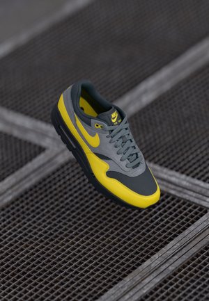 Scarpa Nike Air Max con una toma grigia, accenti gialli e una suola nera texturizzata. Include lacci e dettaglio del logo sulla linguetta.