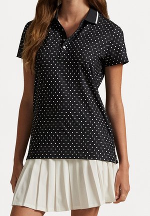 Vrouw met een zwarte korte mouwen poloshirt met witte stippen en een witte geplooide rok tegen een effen achtergrond.