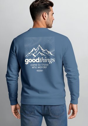Mann trägt blauen Pullover mit Berg-Design und Text "good things come to those who wander" auf dem Rücken, blickt von der Kamera weg.