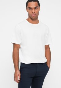 T-shirt bianca a maniche corte con scollo rotondo, in una vestibilità casual, realizzata in morbido cotone. Abbinata a pantaloni blu scuro, indossati con le mani in tasca.
