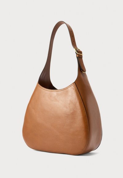 Καφέ δερμάτινη τσάντα ώμου με καμπυλωτή σιλουέτα και ρυθμιζόμενο λουρί με χρυσή αγκράφα, τοποθετημένη σε ανοιχτό φόντο.