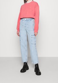 Žena v korálovém zkráceném svetru, světlomodrých vysokých cargo džínách a černých kotníkových botách stojící na neutrálním pozadí.