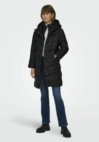 Doudoune noire avec capuche, présentant un design matelassé, fermeture à boutons-pression et poches latérales, associée à un jean bleu et des bottes noires.