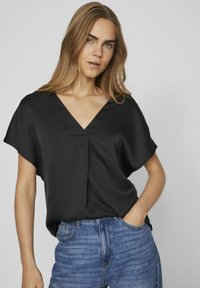 Blusa nera in satin con scollatura a V e maniche corte, caratterizzata da una vestibilità morbida e dettagli di piega discreti, abbinata a jeans in denim blu.