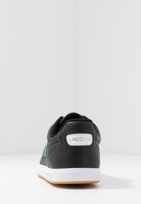 Zapatilla de cuero negro con detalles verdes, paneles laterales texturizados, suela blanca y logo de Lacoste en la parte trasera. Diseño sencillo y de perfil bajo.