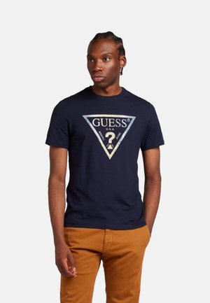 Camiseta de algodón azul marino con un gran gráfico metálico en forma de triángulo que presenta "GUESS" y un signo de interrogación. Mangas cortas, cuello redondo.