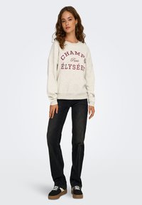 Grå sweatshirt med "CHAMPS ÉLYSÉES" tryckt i vinröd, tillsammans med svarta jeans och svarta sneakers med en beige detalj.