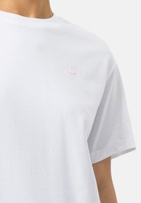 Wit katoenen T-shirt met korte mouwen. Heeft een kleine roze smiley geborduurd op het linkerborstgebied. Gladde textuur.