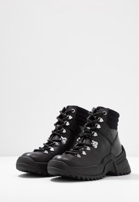 Botas negras altas con acabado de cuero texturizado, con ojales metálicos, suelas gruesas y soporte para el tobillo de punto. Nombre de la marca visible.