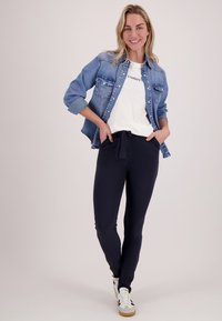 Glimlachende vrouw in een spijkerjack, wit "Couture" T-shirt, zwarte broek met taillekoord en witte sneakers, staand met handen in de zakken.