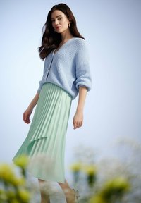 Lichtblauwe gebreide cardigan met een V-hals en oversized mouwen, gecombineerd met een geplooide muntgroene rok, benen zichtbaar, staande pose.