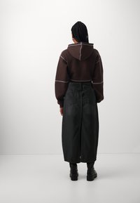 Sudadera corta marrón con costuras blancas en contraste, combinada con una falda negra larga con una abertura trasera y botas negras hasta el tobillo.