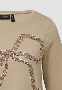 Beige lange mouwtop met een groot, gebogen ontwerp van reflecterende gouden en zilveren pailletten. Ronde hals met een gladde textuur.