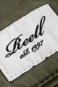 Weißes Stoffetikett mit "Reell" in kursiver schwarzer Schrift und "est. 1997" darunter, auf olivgrünem Stoff mit sichtbarer Naht genäht.