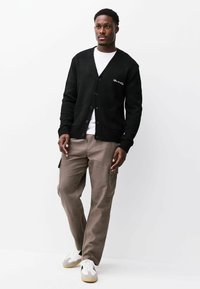 Zwarte cardigan met een V-hals, knoopsluiting en zijzakken, gedragen over een wit shirt en gecombineerd met kaki cargobroek en sneakers.