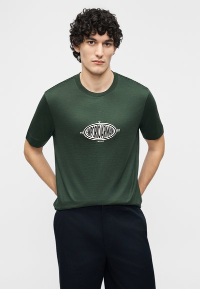 Νέος άντρας με κατσαρά μαλλιά, ντυμένος με σκούρο πράσινο κοντομάνικο Emporio Armani και σκούρο παντελόνι, στέκεται με τα χέρια πίσω από την πλάτη σε γκρι φόντο.