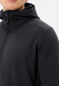 Felpa nera con zip, colletto alto, texture a coste e sottile dettaglio del logo sul petto. Presenta un cappuccio con cordino.