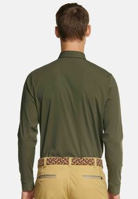 Langärmliges olivgrünes Hemd mit Kragen, kombiniert mit hellen khakifarbenen Hosen und einem gewebten mehrfarbigen Gürtel mit Zickzackmuster.