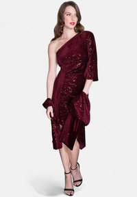 Robe bordeaux à une épaule ornée de sequins, avec une coupe ajustée, une manche drapée et un détail de fente sur le côté, accompagnée de talons noirs.