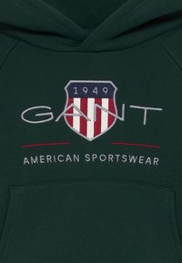 Sudadera verde oscuro con un logo de escudo que tiene franjas rojas y blancas, "GANT" en bordado gris, y "1949" arriba con "AMERICAN SPORTSWEAR" debajo.