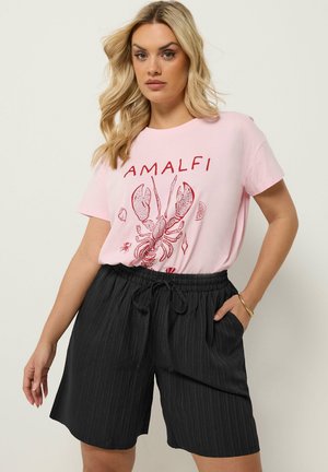 Blondynka w różowym t-shircie z czerwonym homarem i napisem "AMALFI", zestawiona z czarnymi szortami ze sznurkiem i złotą bransoletką.
