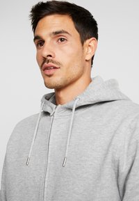 Grijze zip-up hoodie gemaakt van zachte stof met een trekkoordcapuchon en metalen accenten op de rits, met een gladde textuur en een casual pasvorm.
