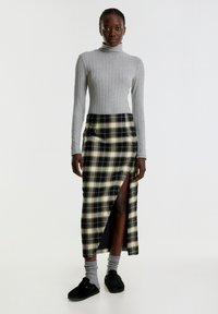 Grau gerippter Rollkragenpullover und schwarzer Maxi-Rock mit kariertem Muster und gelben Akzenten; der Rock hat einen vorderen Schlitz. Modell trägt graue Socken und schwarze Schuhe.