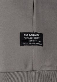 Etichetta tessile grigia con testo nero che legge "KEY LARGO", con ulteriori dettagli di branding, cucita su una superficie di tessuto liscia.