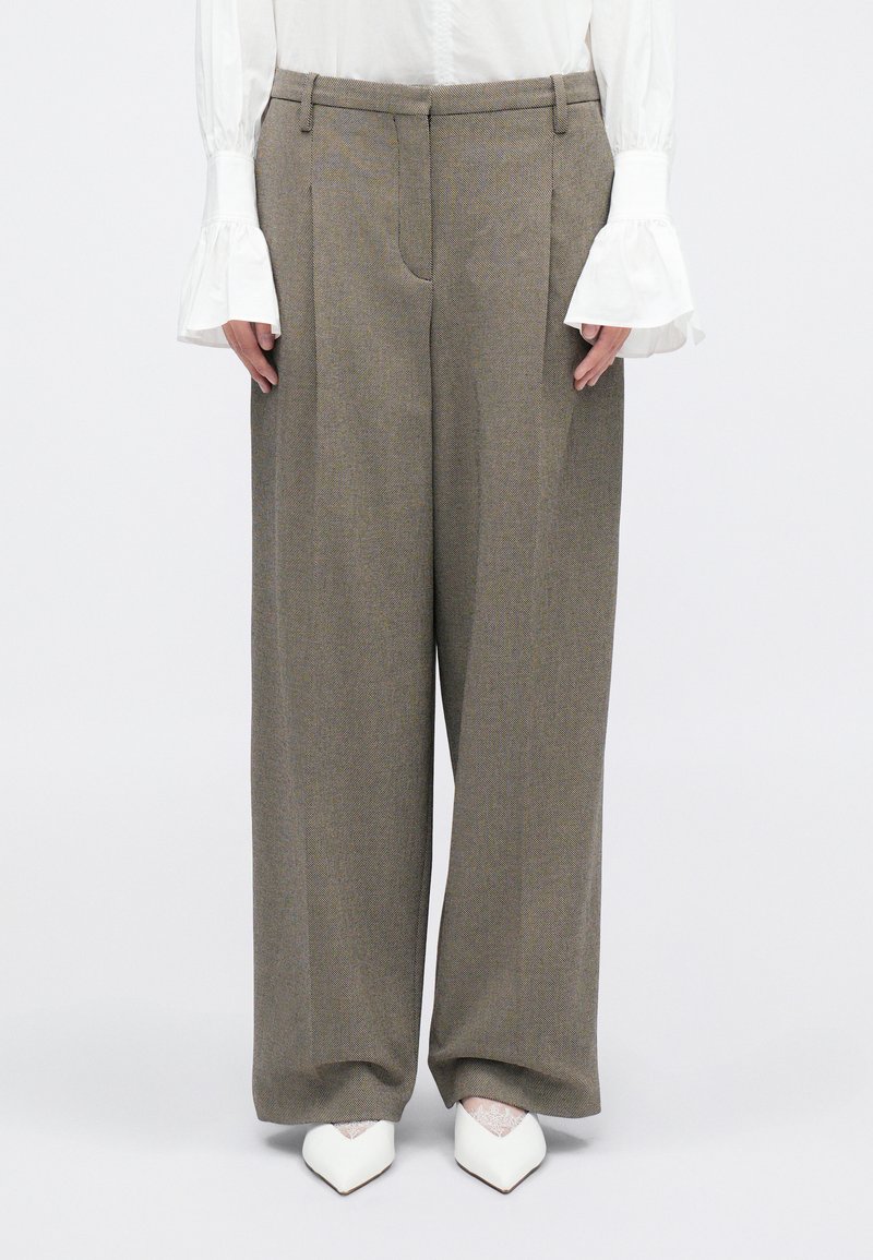 Pantalons à jambes larges en tissu marron texturé, dotés d'une taille haute, de plis avant et de poches latérales. Associés à des chaussures pointues blanches.