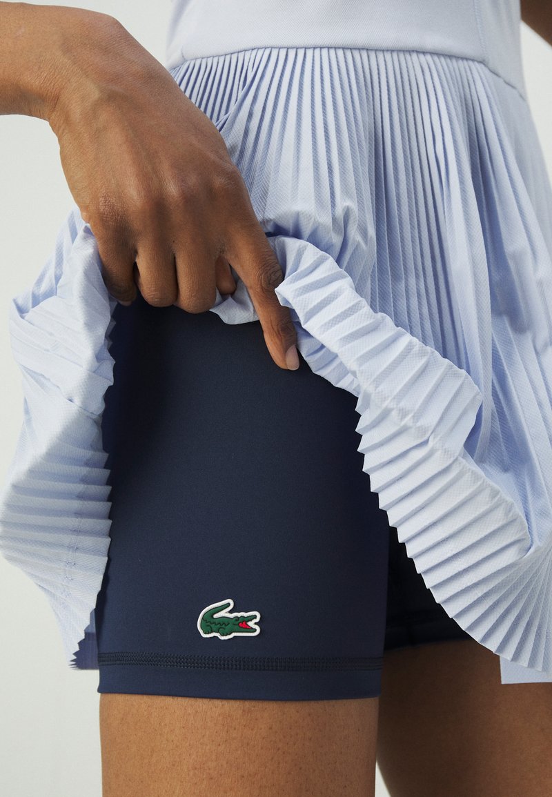 Marinefarbene Sportshorts mit einer gefalteten, hellblauen Überlagerung. Mit Logo-Aufnäher. Die Hand hält die Falten und zeigt die weiche Textur der Shorts.